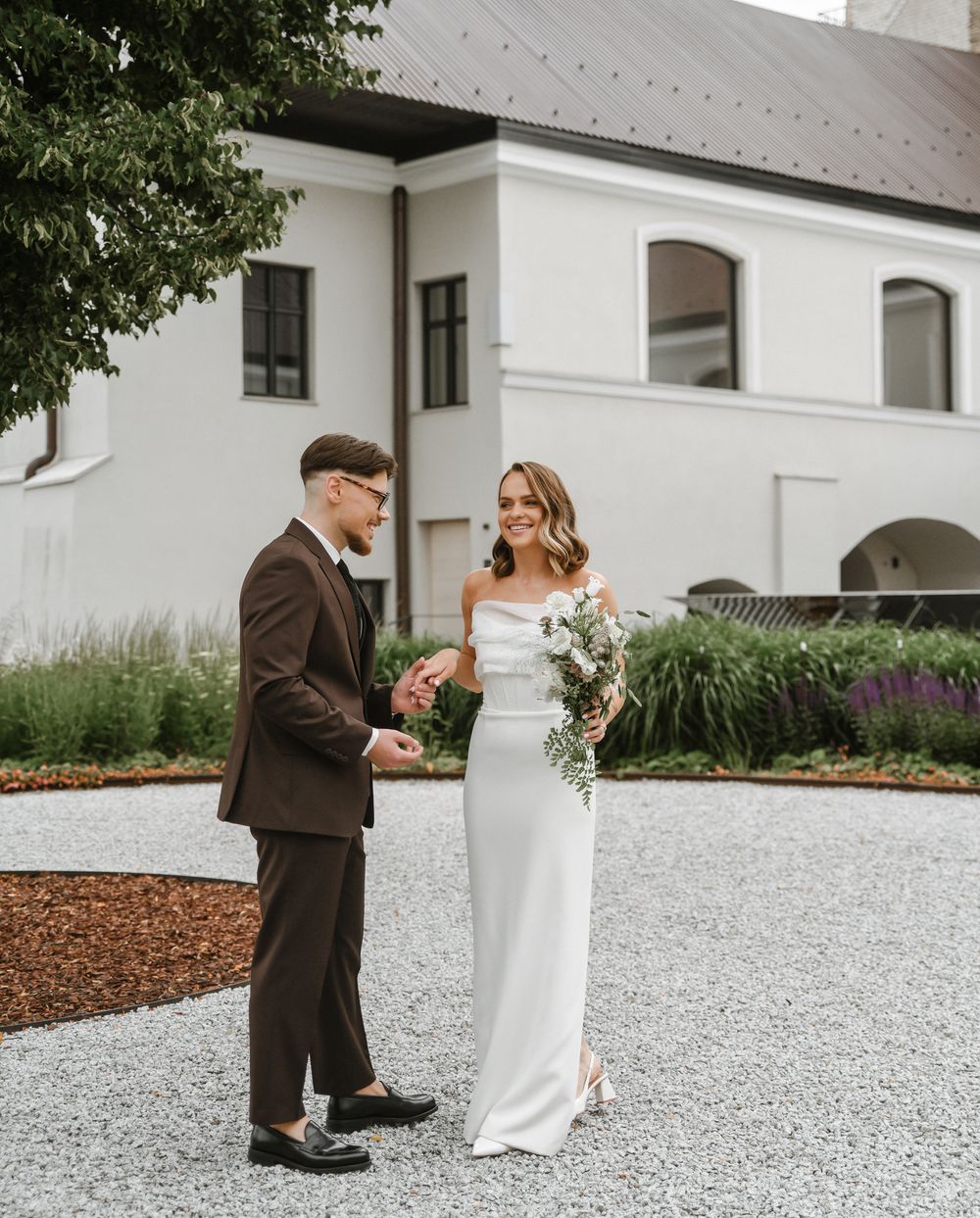 Elegant couple stands on white gravel path in front of white church building for outdoor wedding photo.
