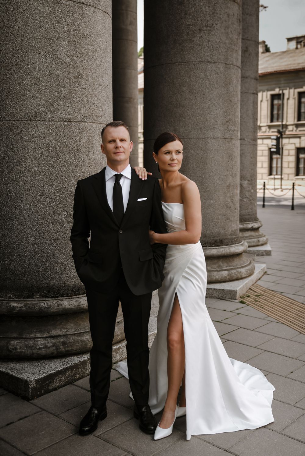 Elegant wedding portrait between classical stone columns with dramatic architectural backdrop.
