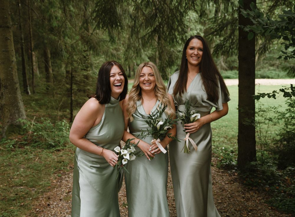 A group of bridesmaids in sage green dresses share candid laughing moments while walking through a wooded forest path.