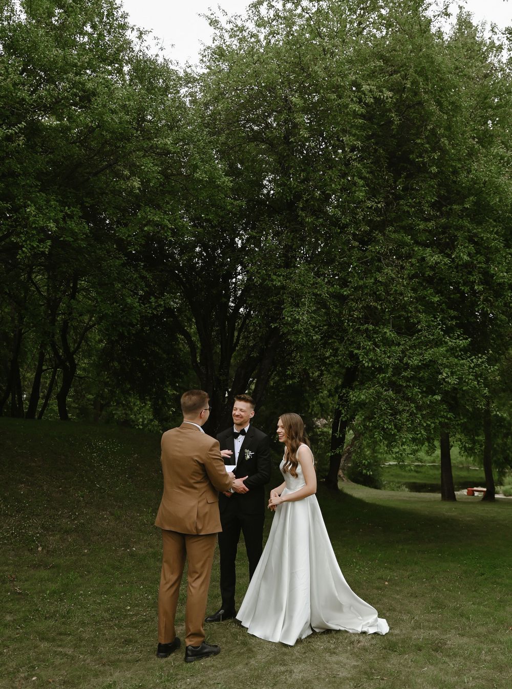 A couple shares a romantic moment on a grassy hillside surrounded by lush green trees during an outdoor photo session.