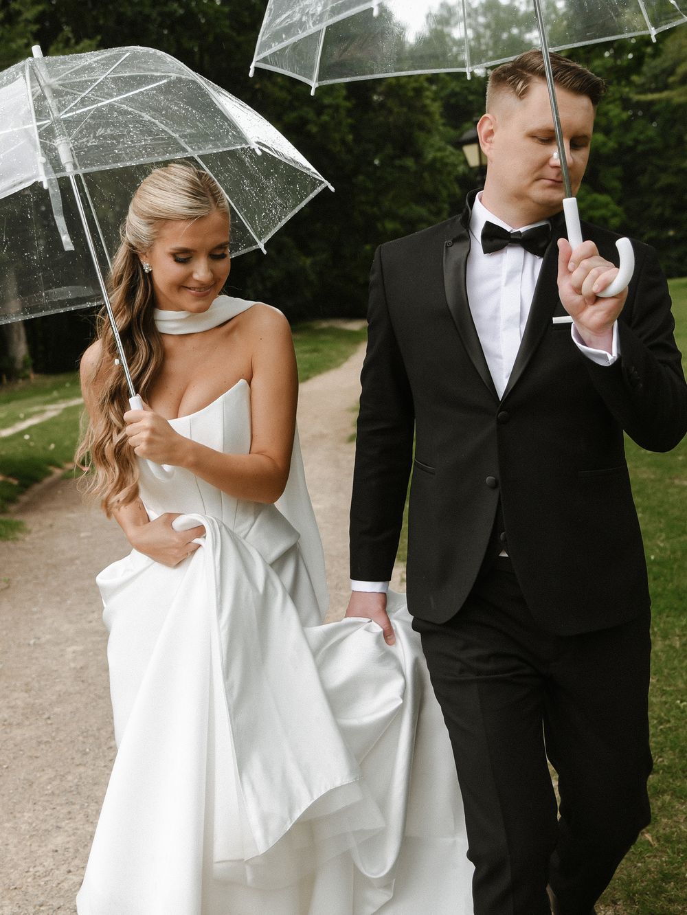A couple on their wedding day sharing a playful moment under clear umbrellas on a path surrounded by greenery.
