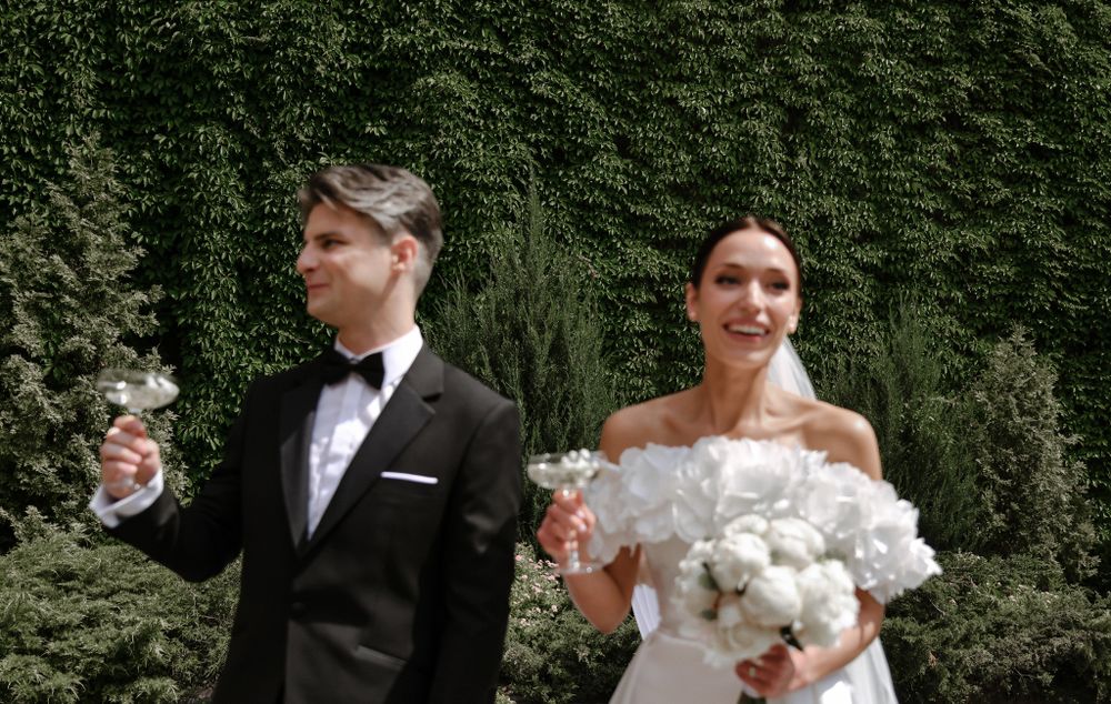 A couple poses together outdoors against greenery, with the bride holding a white bouquet and groom holding a glass.