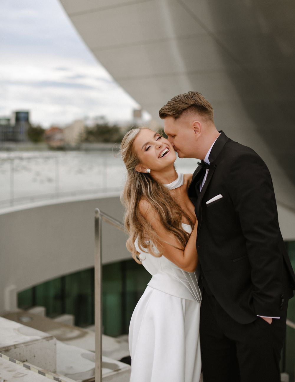 A bride and groom share intimate romantic moments under a concrete structure in a stylish modern wedding photoshoot.