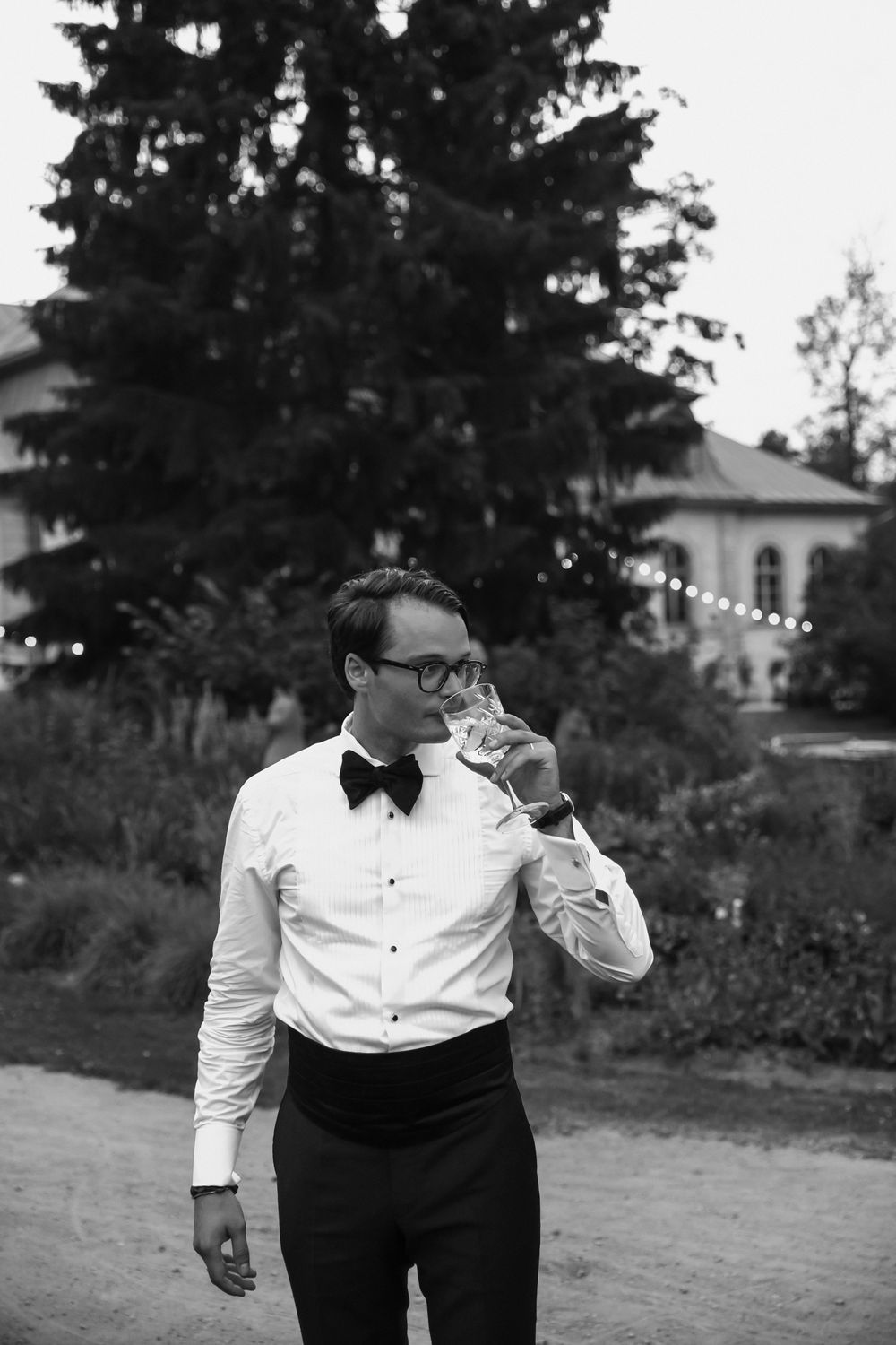 A groom in formal attire sips champagne outdoors during a wedding celebration.