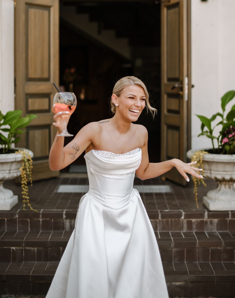 Bride in elegant white satin dress laughing and dancing joyfully on a porch with brick columns.