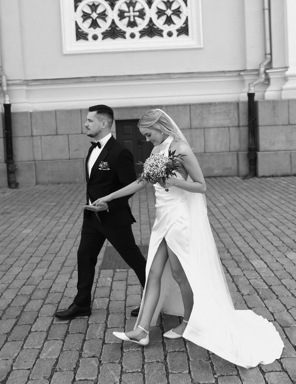A bride in a flowing white gown with a slit and groom in a black tuxedo walk together on cobblestone pavement.