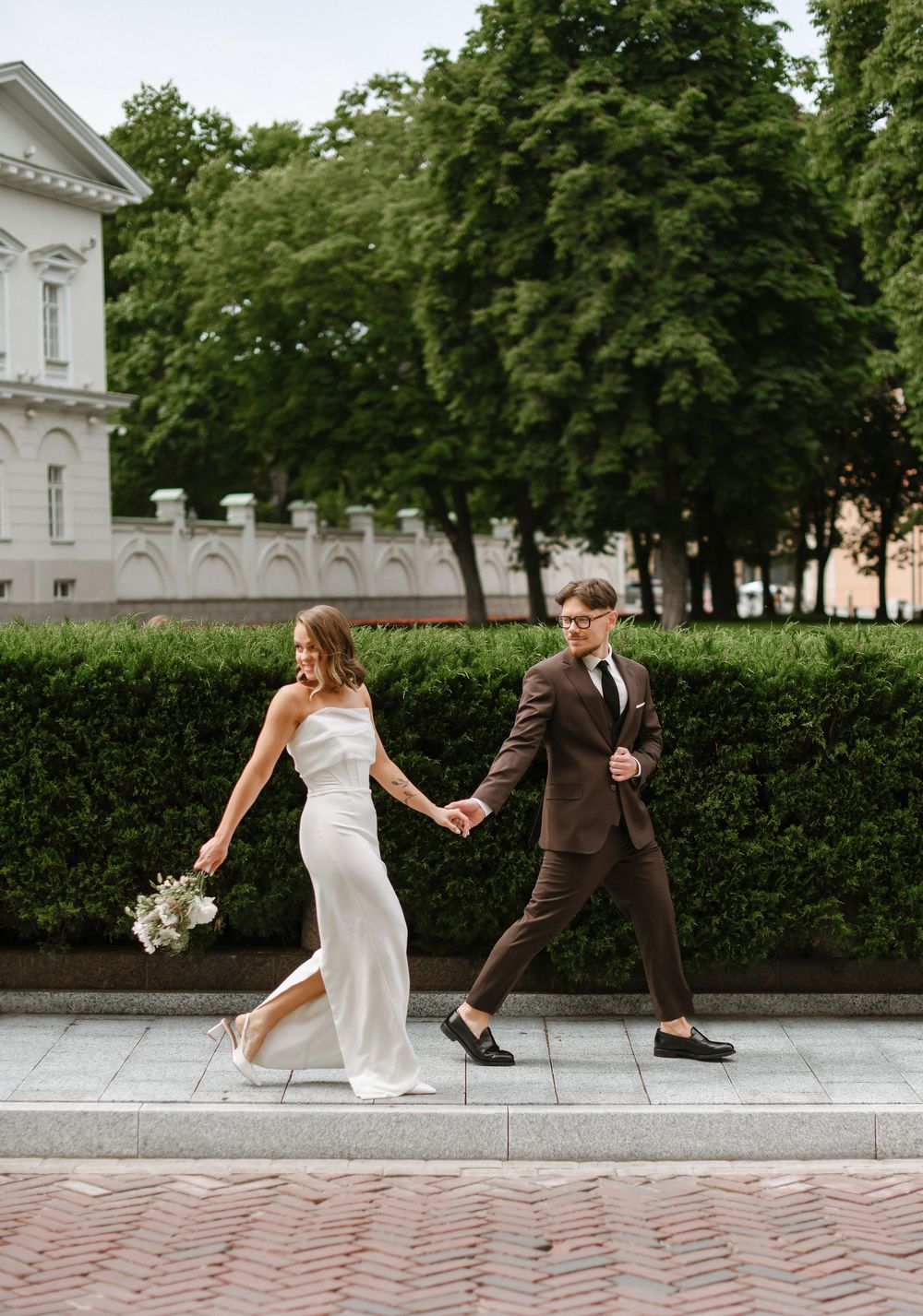 A couple in wedding attire dances playfully on a stone pathway near manicured hedges and historic architecture.