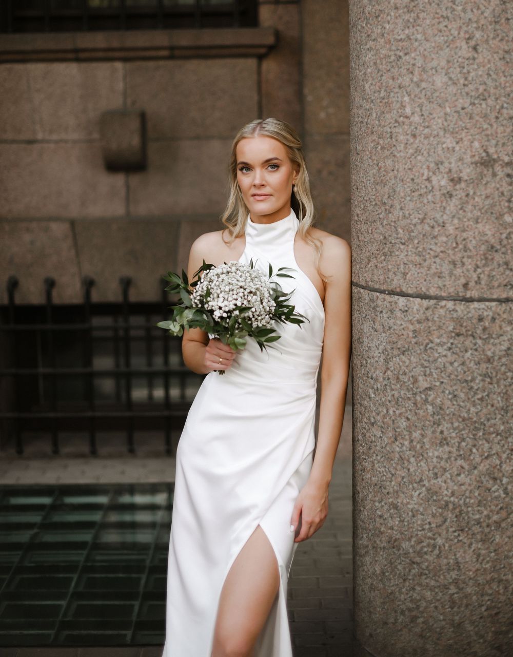 Elegant bride in a sleek white wedding gown with a high slit poses gracefully against a stone column backdrop.