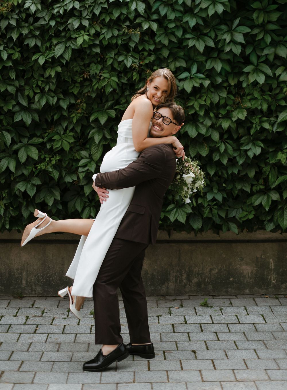 A couple in wedding attire shares a tender moment against a green vine-covered wall background.