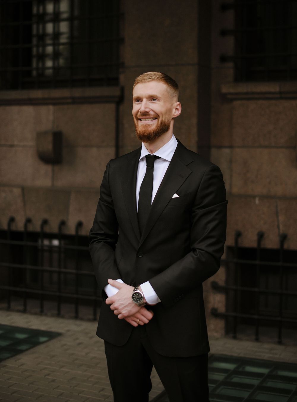 A groom in a classic black suit and tie stands outside a historic building with a joyful expression.