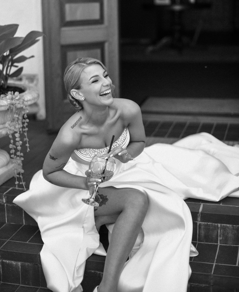 A joyful bride in a strapless white gown sits on steps holding a champagne glass while laughing in black and white.