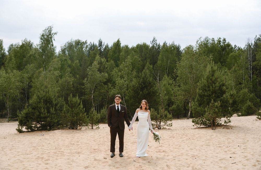 A couple in wedding attire walks together on a sandy beach surrounded by evergreen trees.