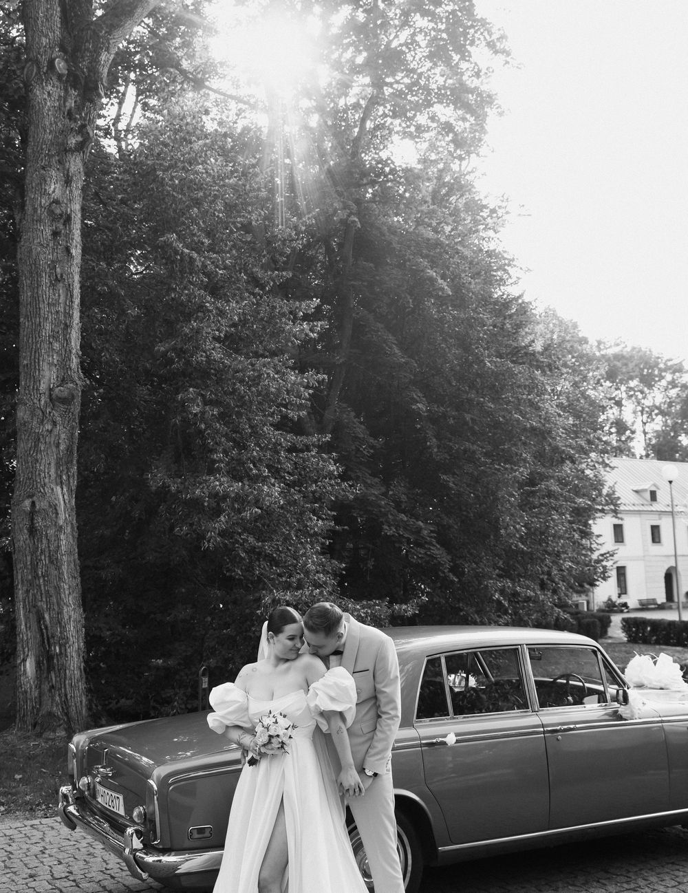 A classic black vintage car parked among trees serves as an elegant backdrop for a wedding celebration.
