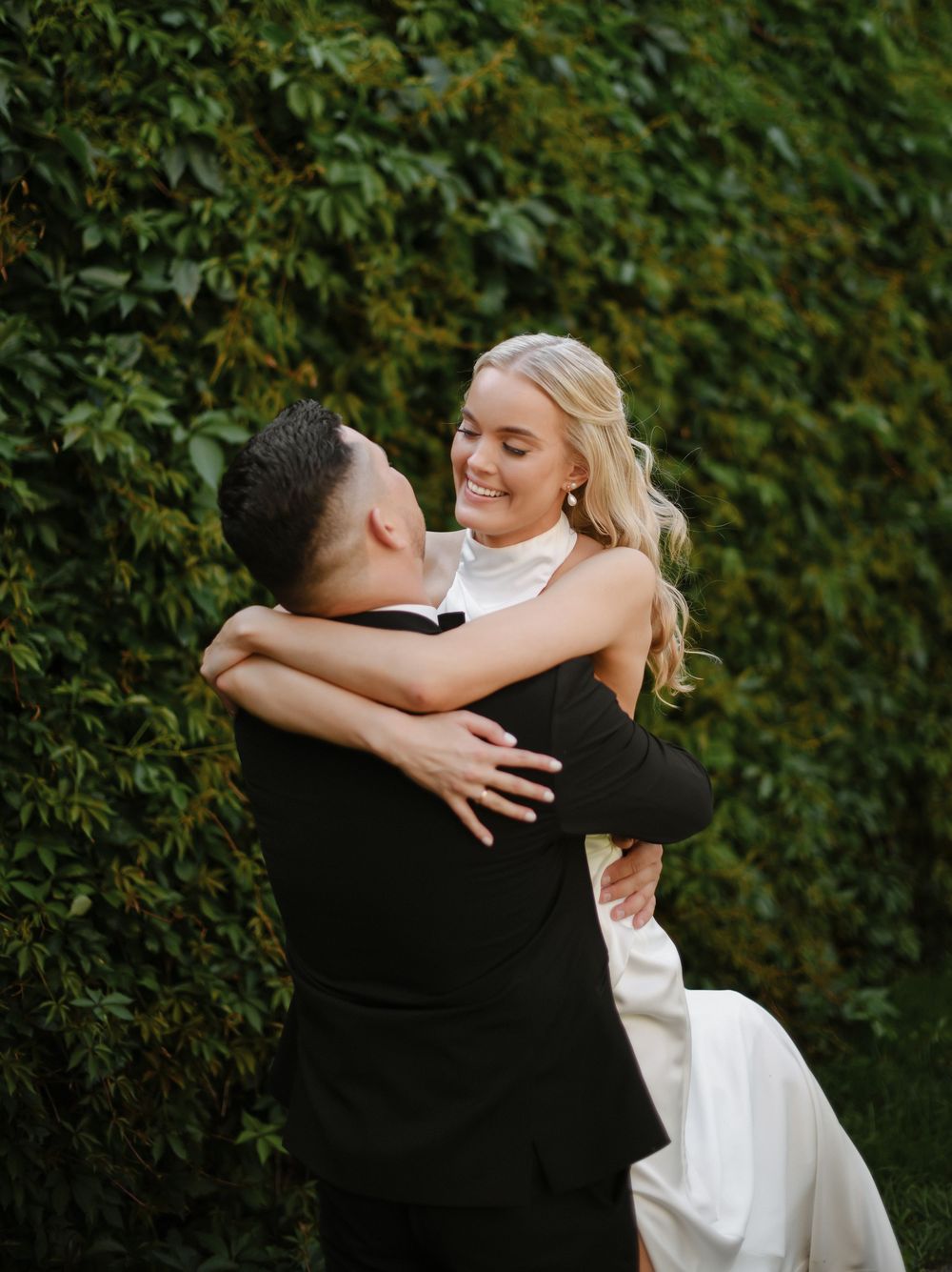 Couple shares intimate embrace outdoors against lush green ivy wall during wedding photoshoot.