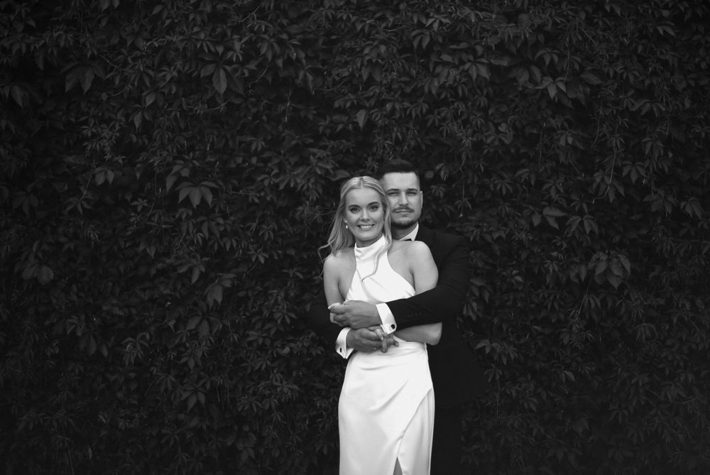 Black and white portrait of a couple embracing outdoors against a hedge background.