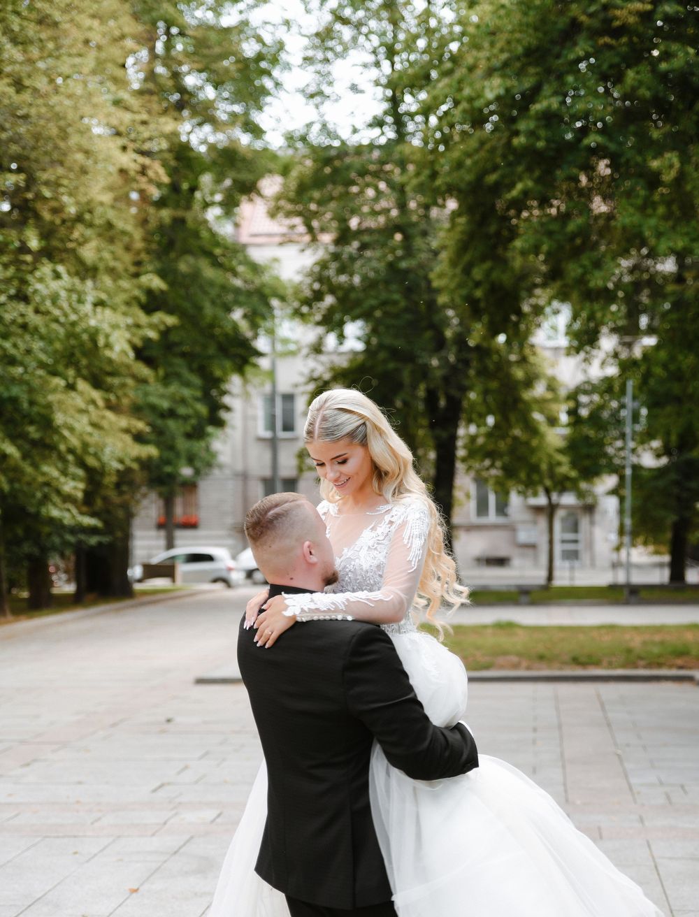 A couple in formal wedding attire dancing and twirling together on a tree-lined pathway.