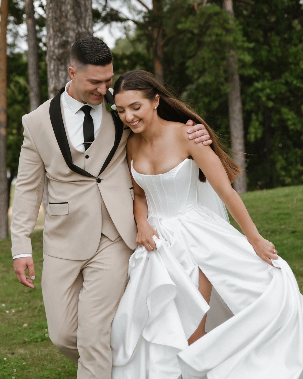 Wedding party walks together outdoors while bride lifts her white dress, with groomsmen wearing beige suits.
