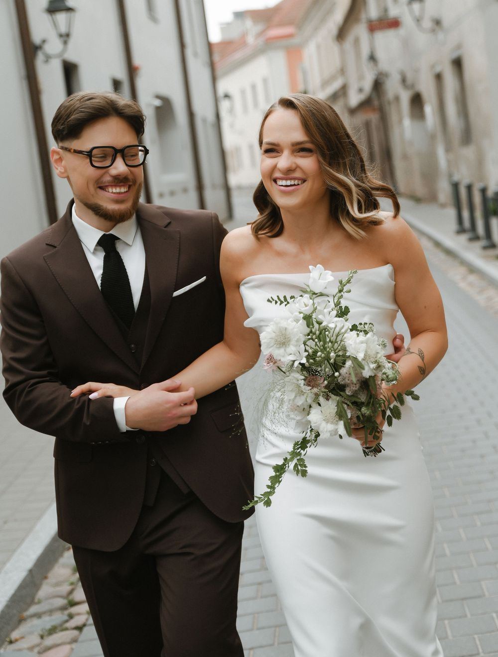 Happy couple in wedding attire share joyful moments walking together on a cobblestone street.