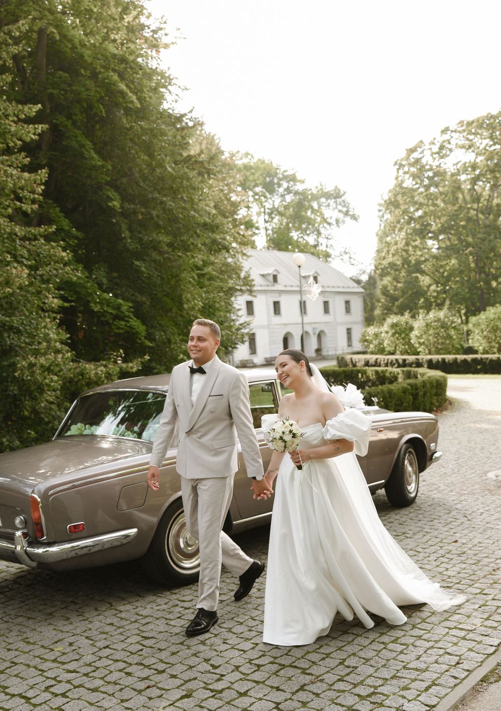 Elegant wedding couple poses with vintage brown luxury car on cobblestone driveway at historic white mansion.