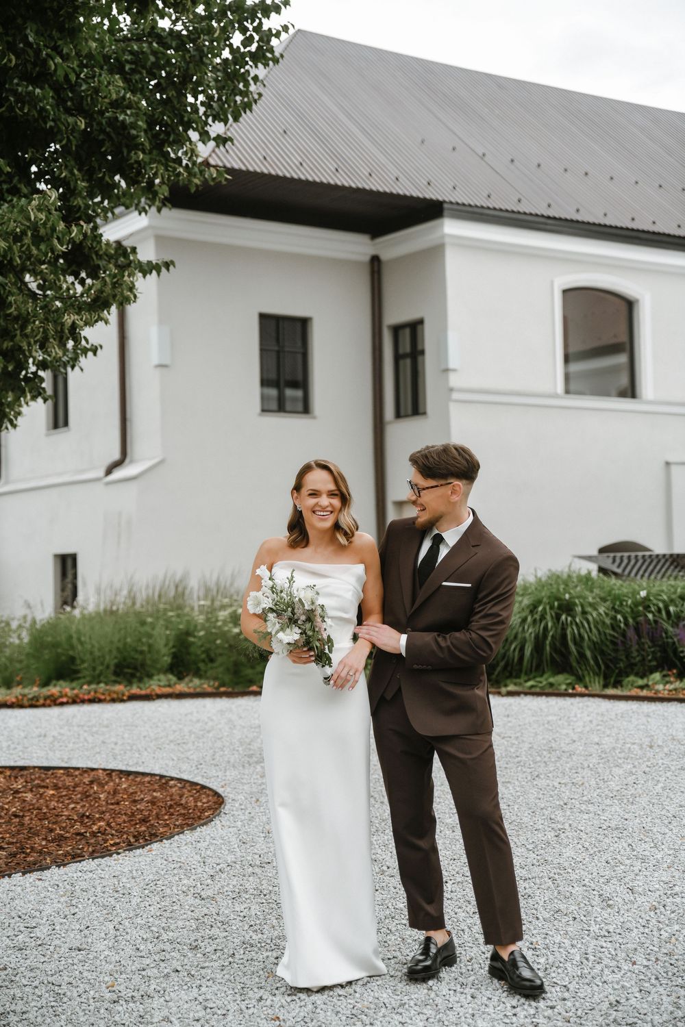 A couple in wedding attire shares a romantic moment on a white gravel path beside a modern white building.
