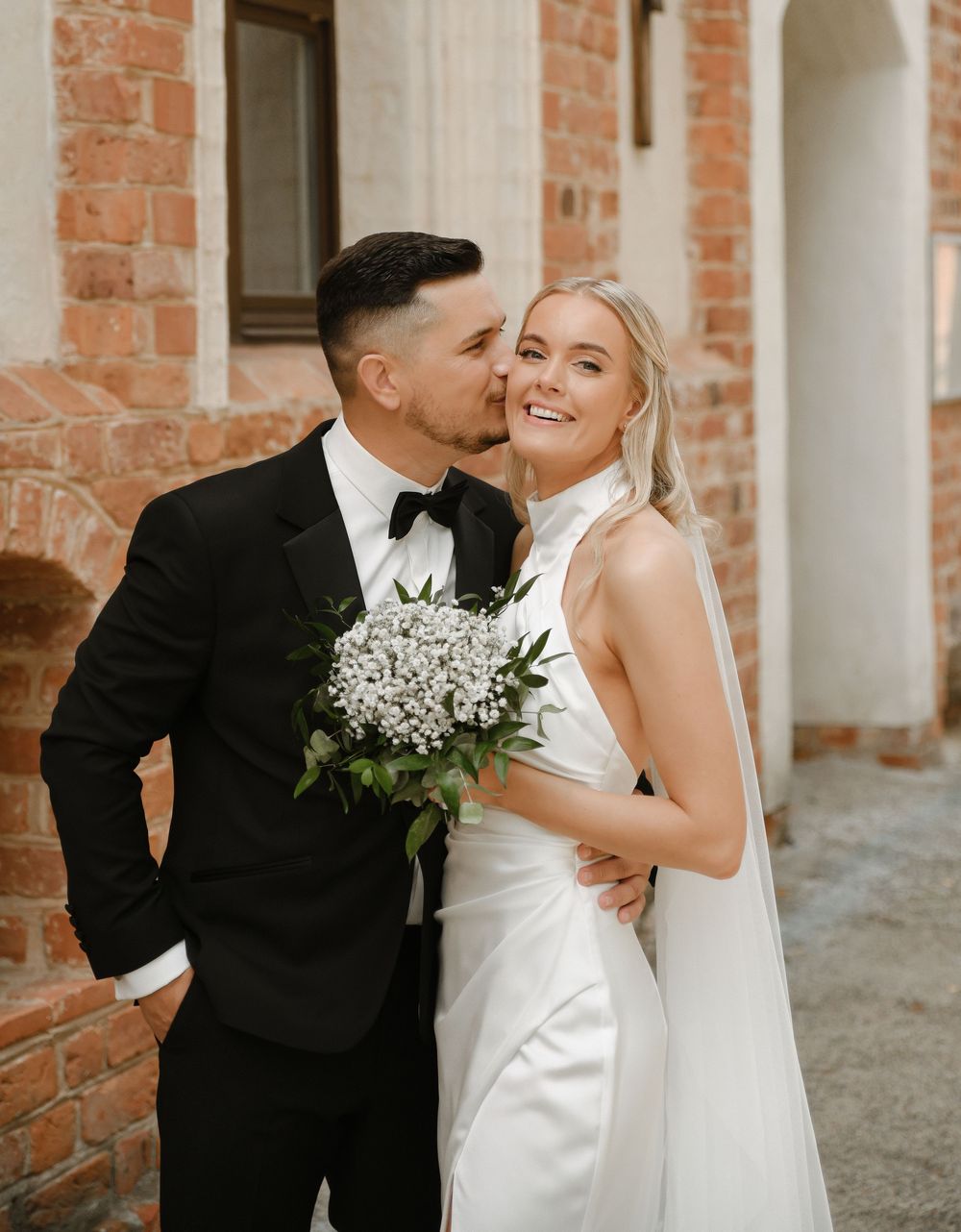 A bride and groom share intimate moments against a rustic brick wall with white columns.