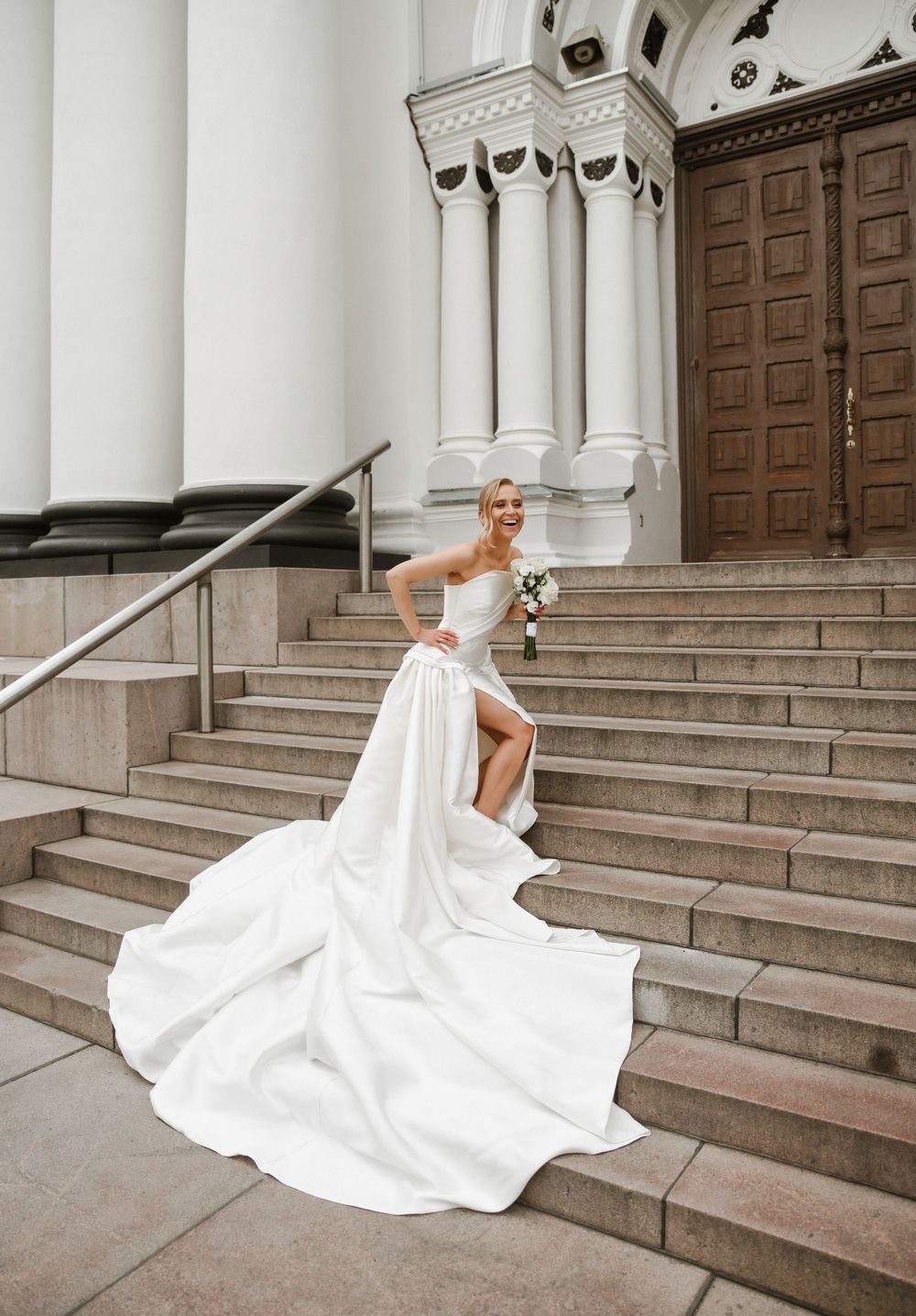 A bride in a flowing white wedding gown gracefully descends stone church steps as her dress billows in the wind.