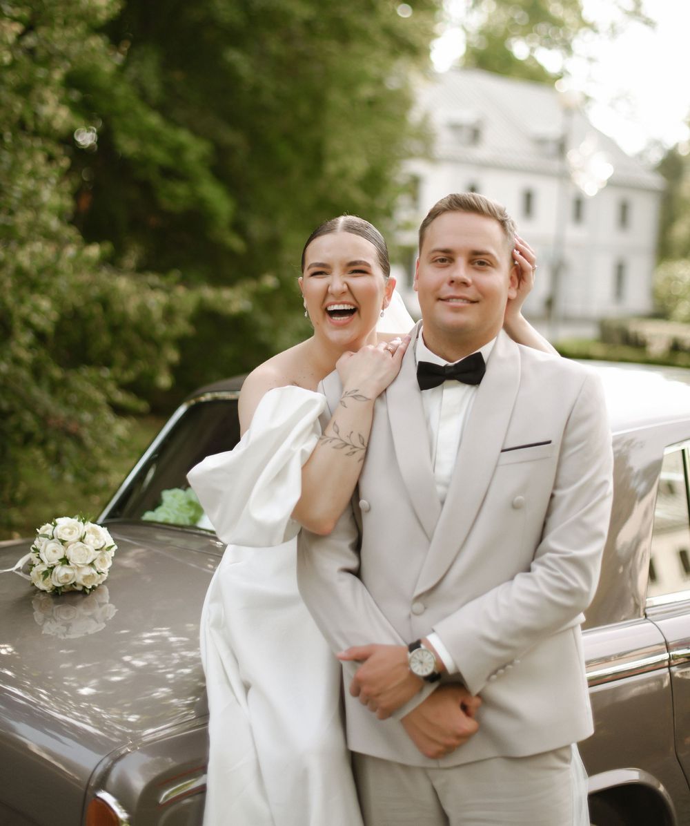 A wedding couple shares joyful candid moments together in front of a vintage car on their special day.