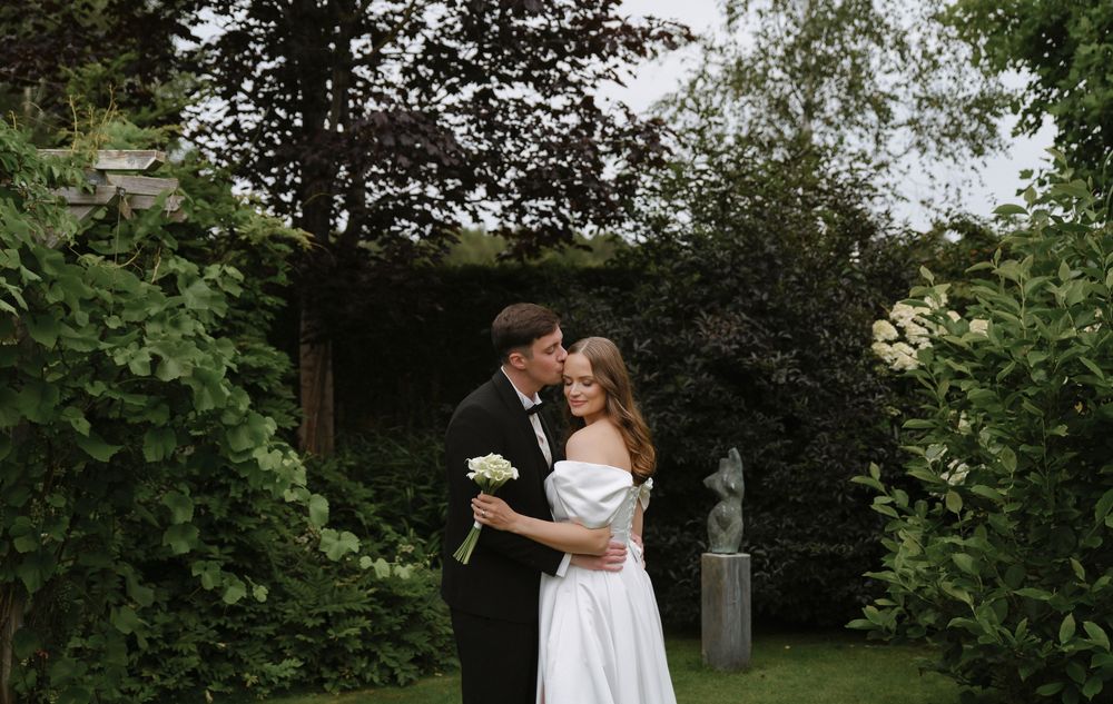 A romantic garden wedding moment with a couple embracing among lush green foliage on a cloudy day.