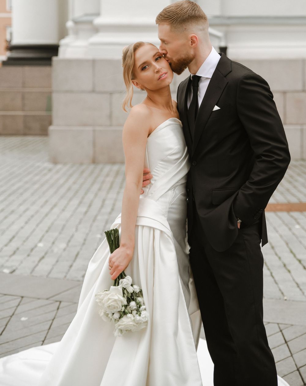 Elegant wedding couple embraces outdoors with bride in white gown holding bouquet and groom in classic black suit.