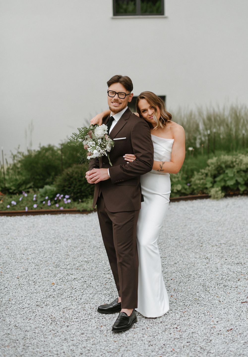 A couple in wedding attire share a tender moment on a gravel path with greenery in the background.
