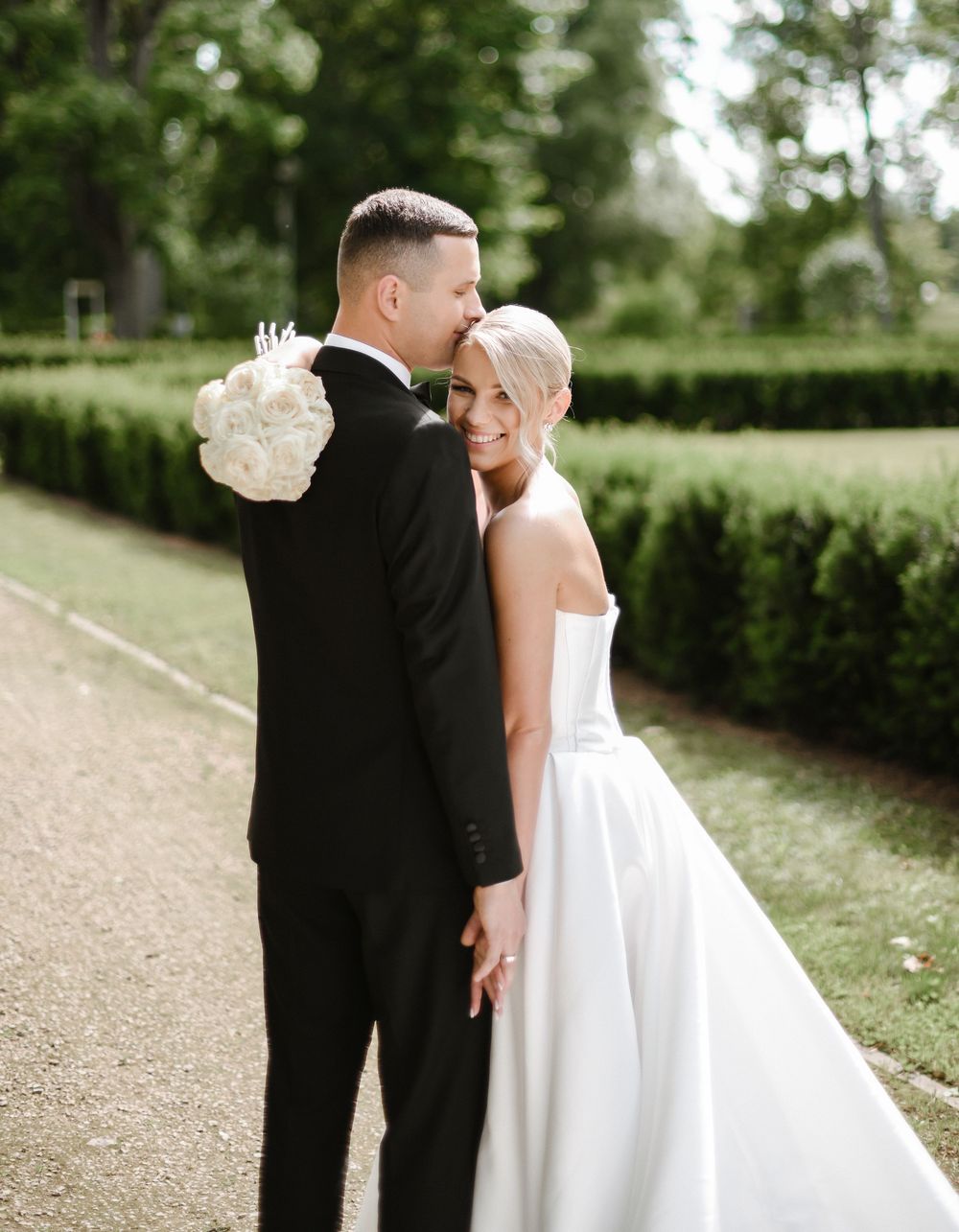 A couple in wedding attire share an intimate moment together on a gravel path surrounded by lush greenery.