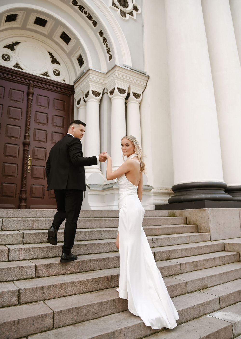 Elegant wedding couple poses on grand stone steps with white columns and wooden doors of historic building.