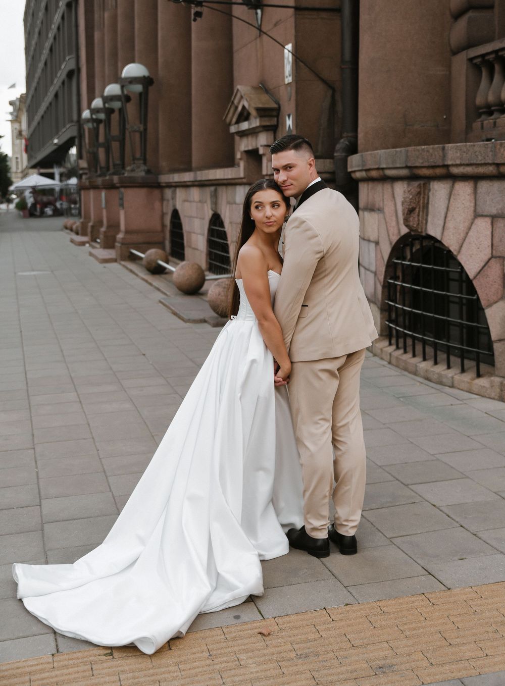 A couple in wedding attire shares an intimate moment against a stone building backdrop on a city sidewalk.