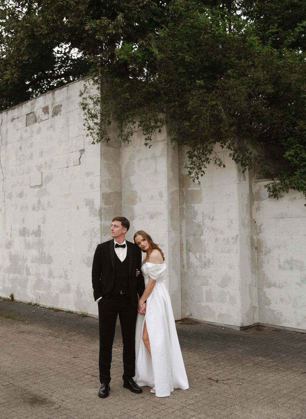 A couple in formal wedding attire stands together against a white stone wall in an outdoor setting.