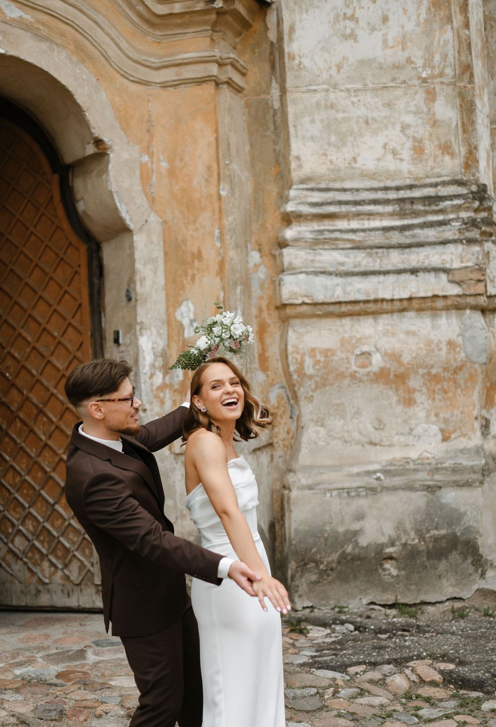 A couple in wedding attire shares a playful moment against a weathered stone wall with rustic wooden doors.