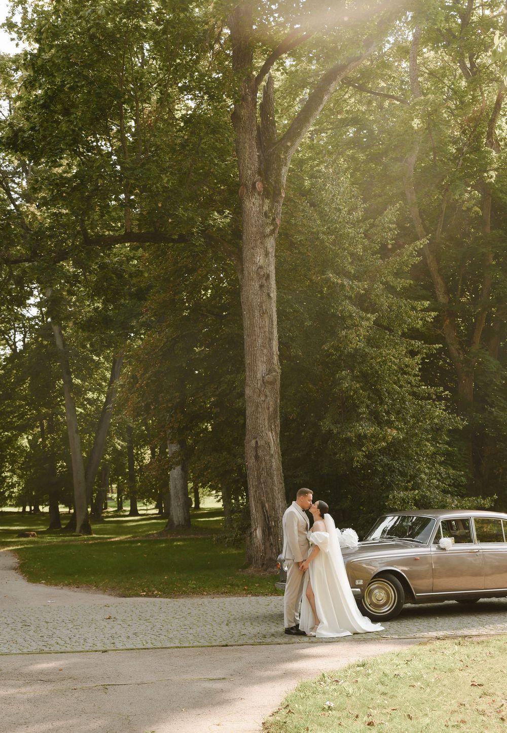 A couple stands beside a vintage car under tall trees in a sunlit park setting with a dreamy, romantic atmosphere.