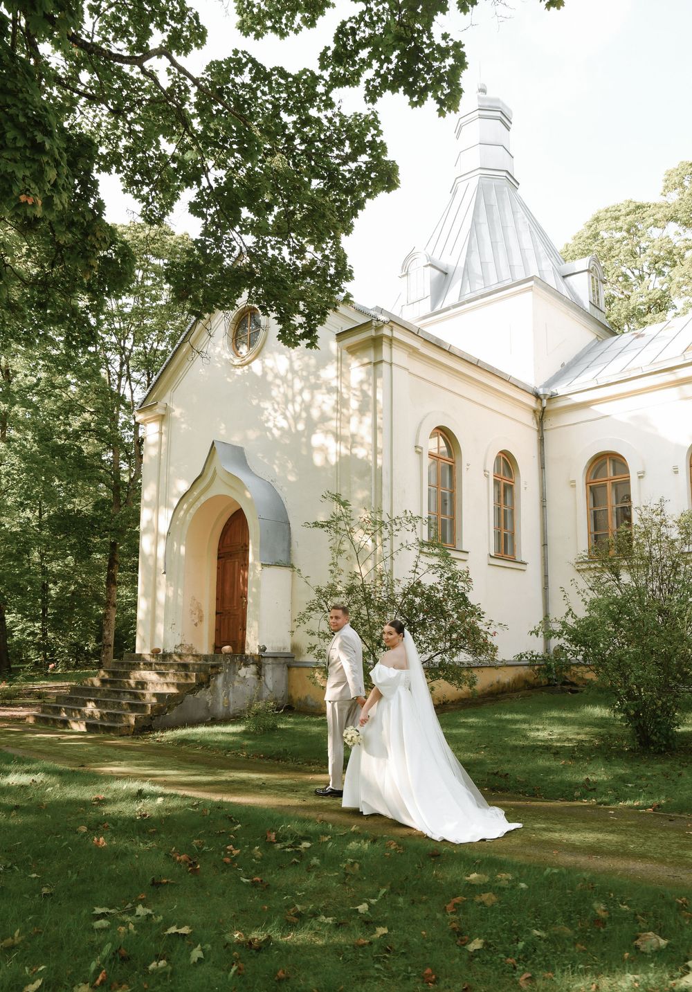 A couple stands outside a beautiful white church with a spired tower surrounded by lush green trees on a sunny day.