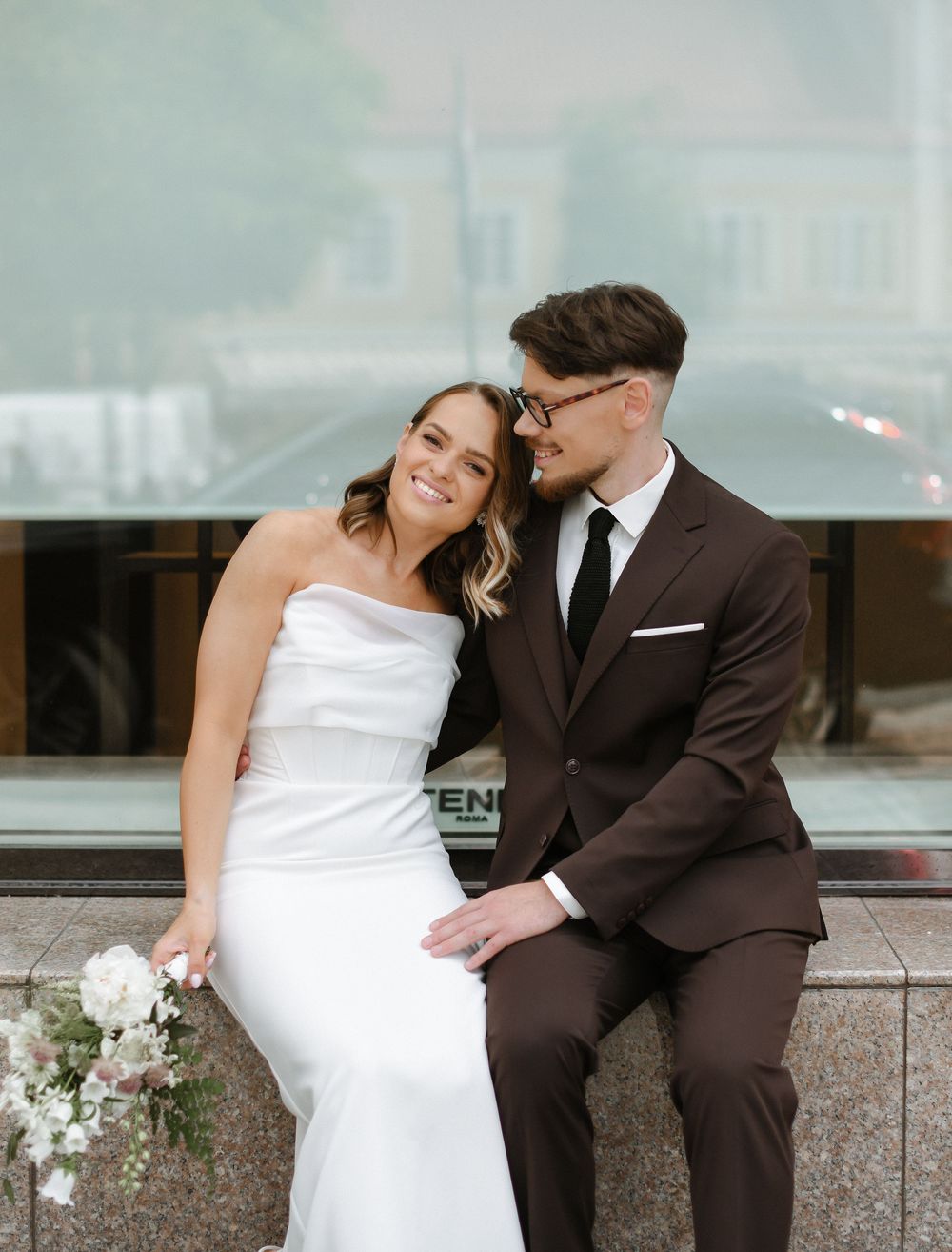 A couple in wedding attire shares a romantic moment while sitting on a stone ledge in an urban setting.