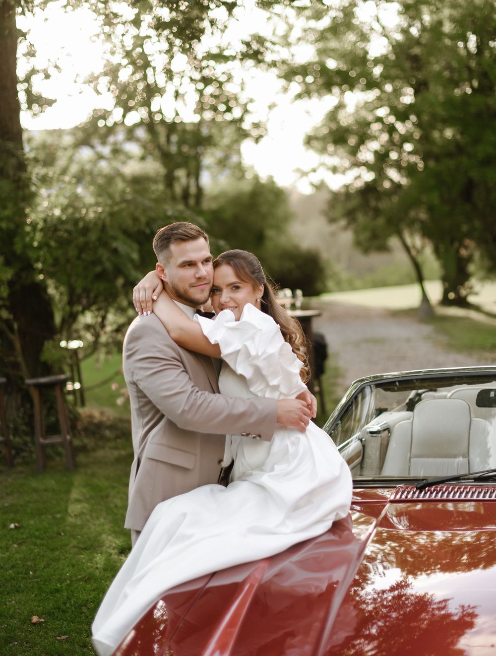 A romantic wedding couple embraces next to a classic red convertible car in a lush garden setting.
