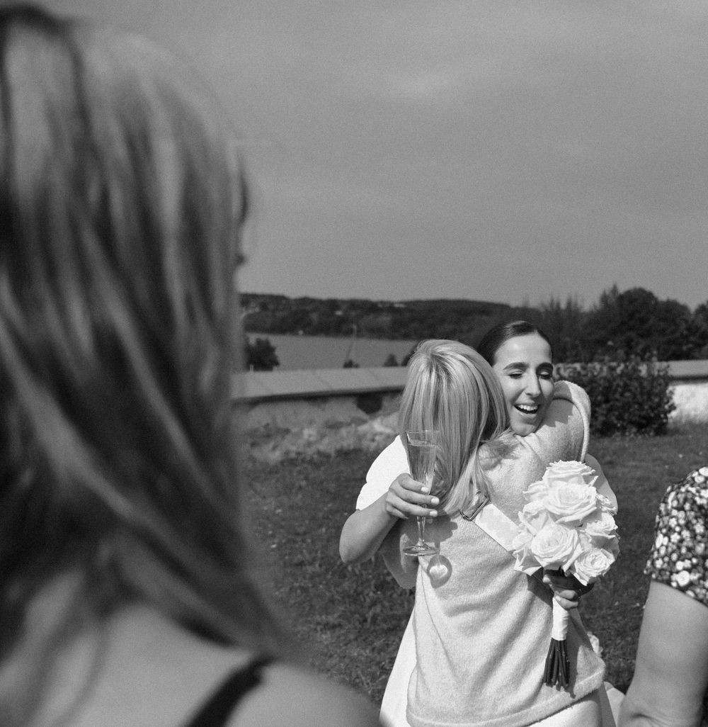 An emotional black and white photo shows wedding guests embracing while holding bouquets outdoors.