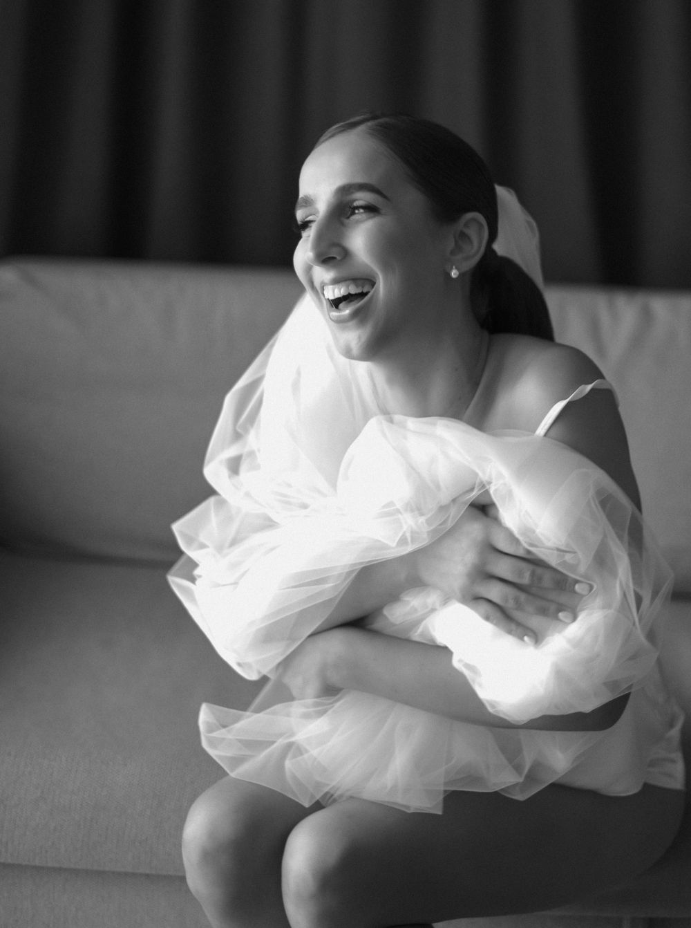 A bride in a white dress hugs a bundle of tulle fabric while sitting on a couch in a black and white photo.