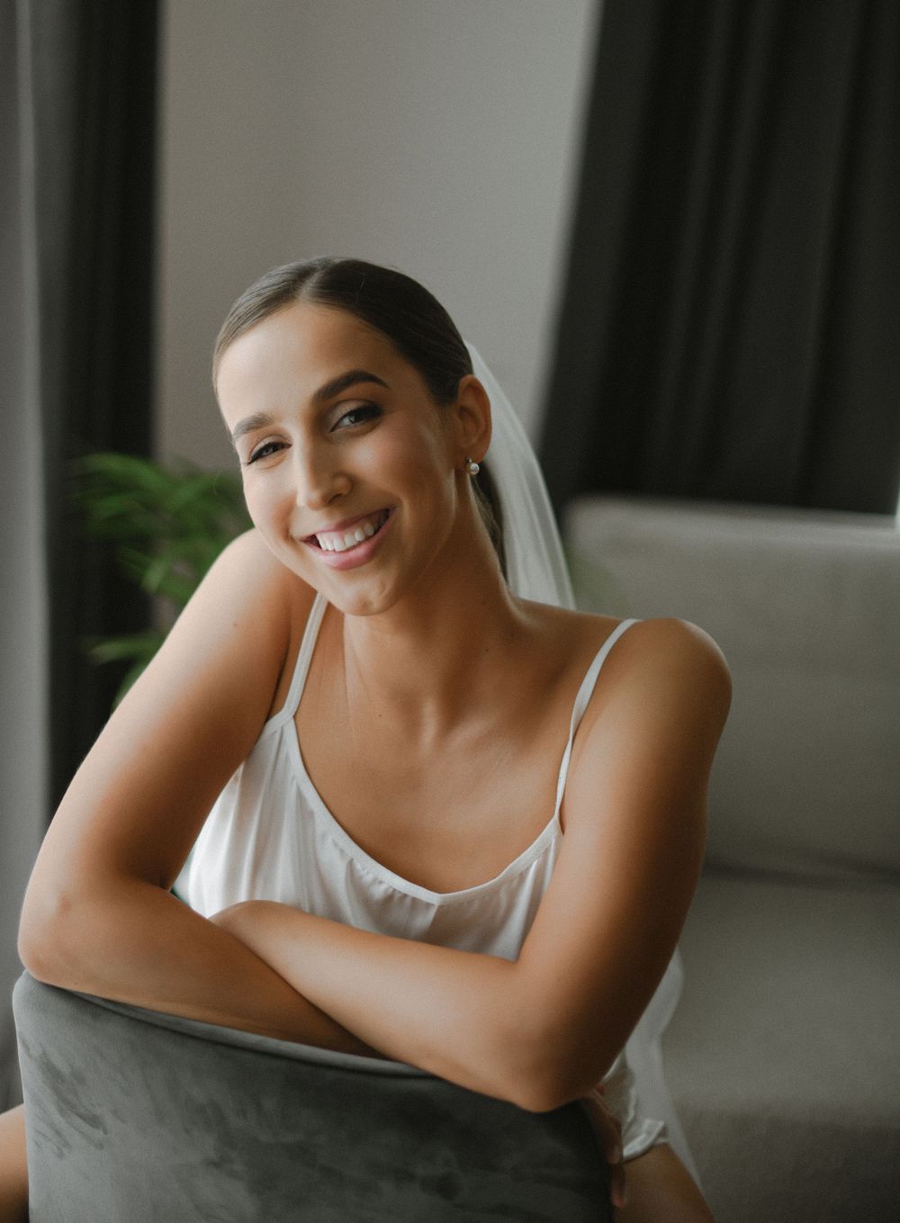 A joyful bride in a white tank top sits casually with arms crossed during wedding preparations.