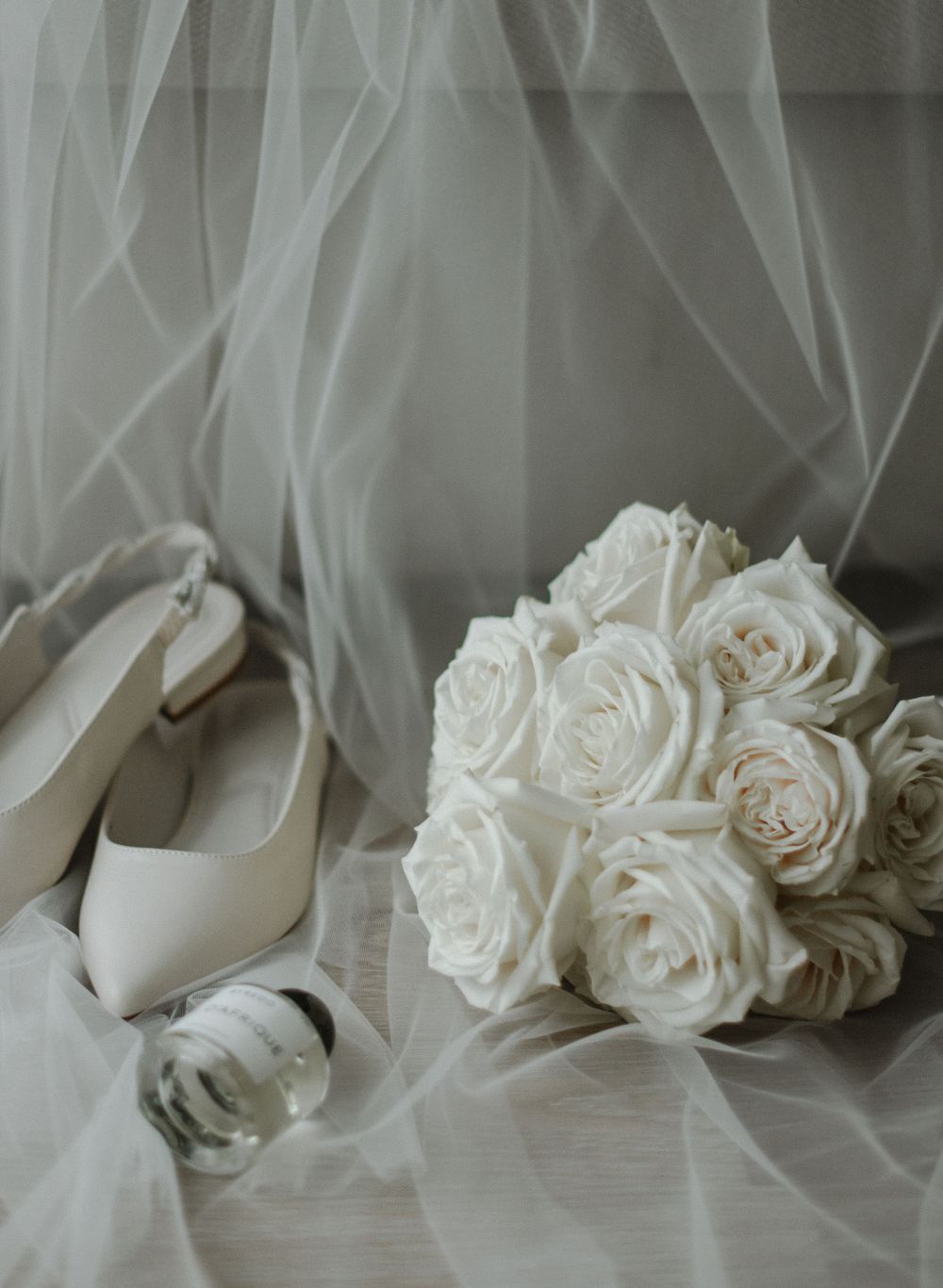 Elegant white roses and bridal shoes arranged with flowing veil on light surface.