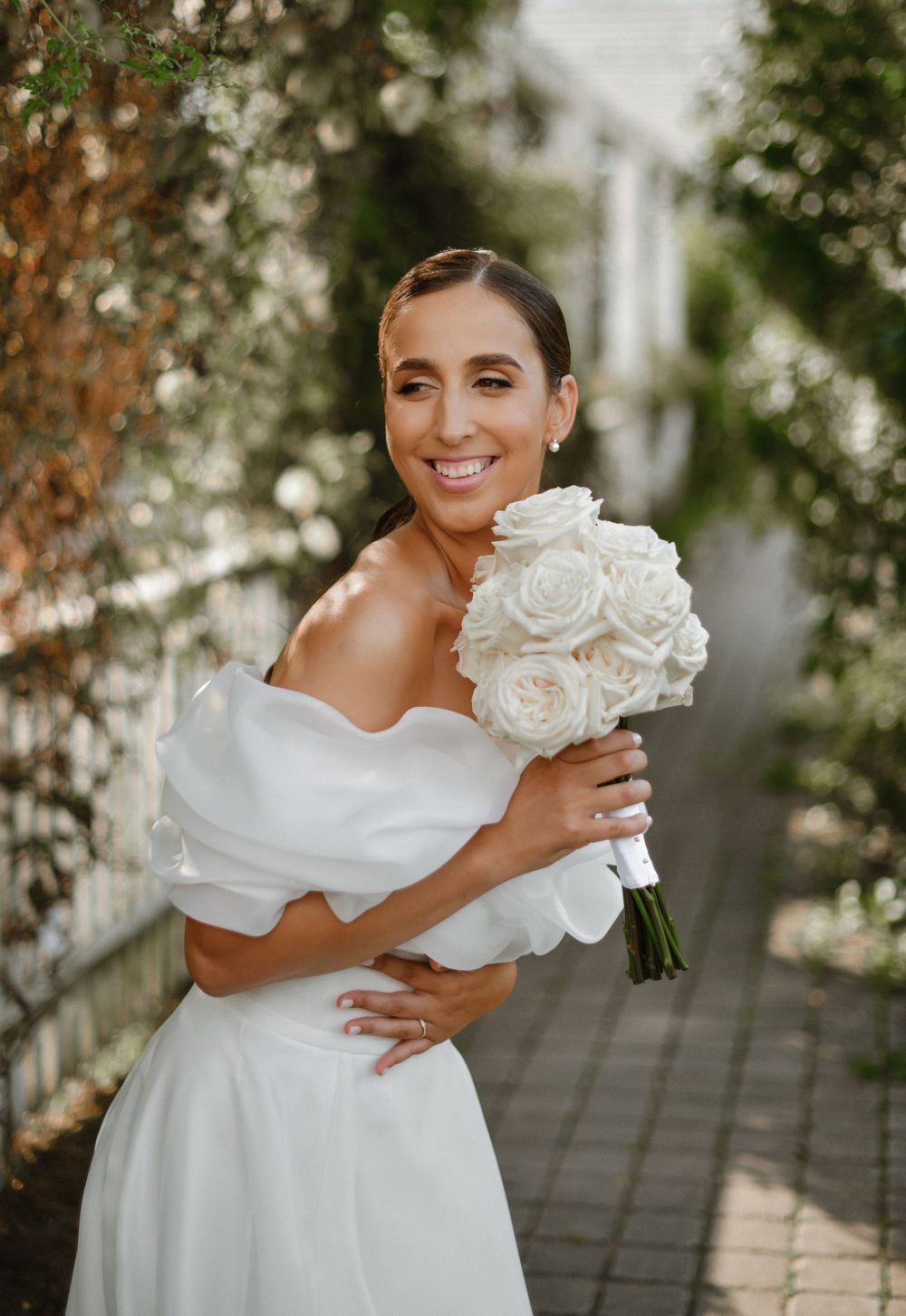 Bride in off-shoulder white gown holds classic white rose bouquet while posing on garden path.