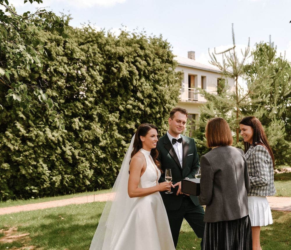 A couple in formal wedding attire stands together in a garden setting during an outdoor ceremony.