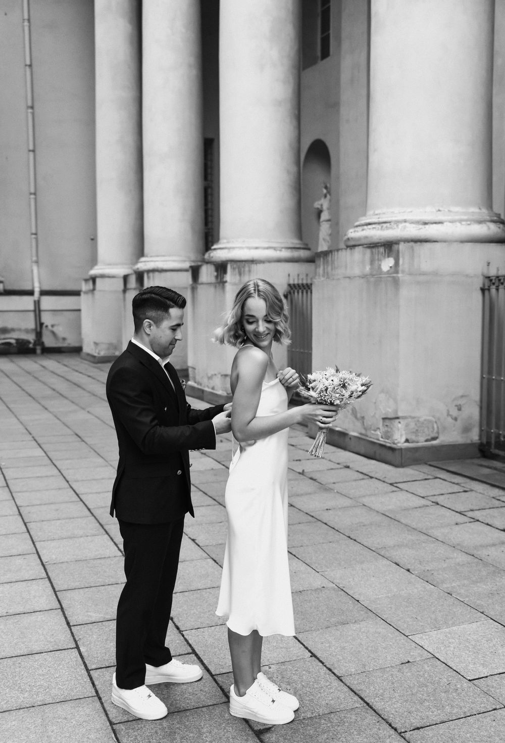 Black and white photo of a couple in wedding attire posing by classical columns.