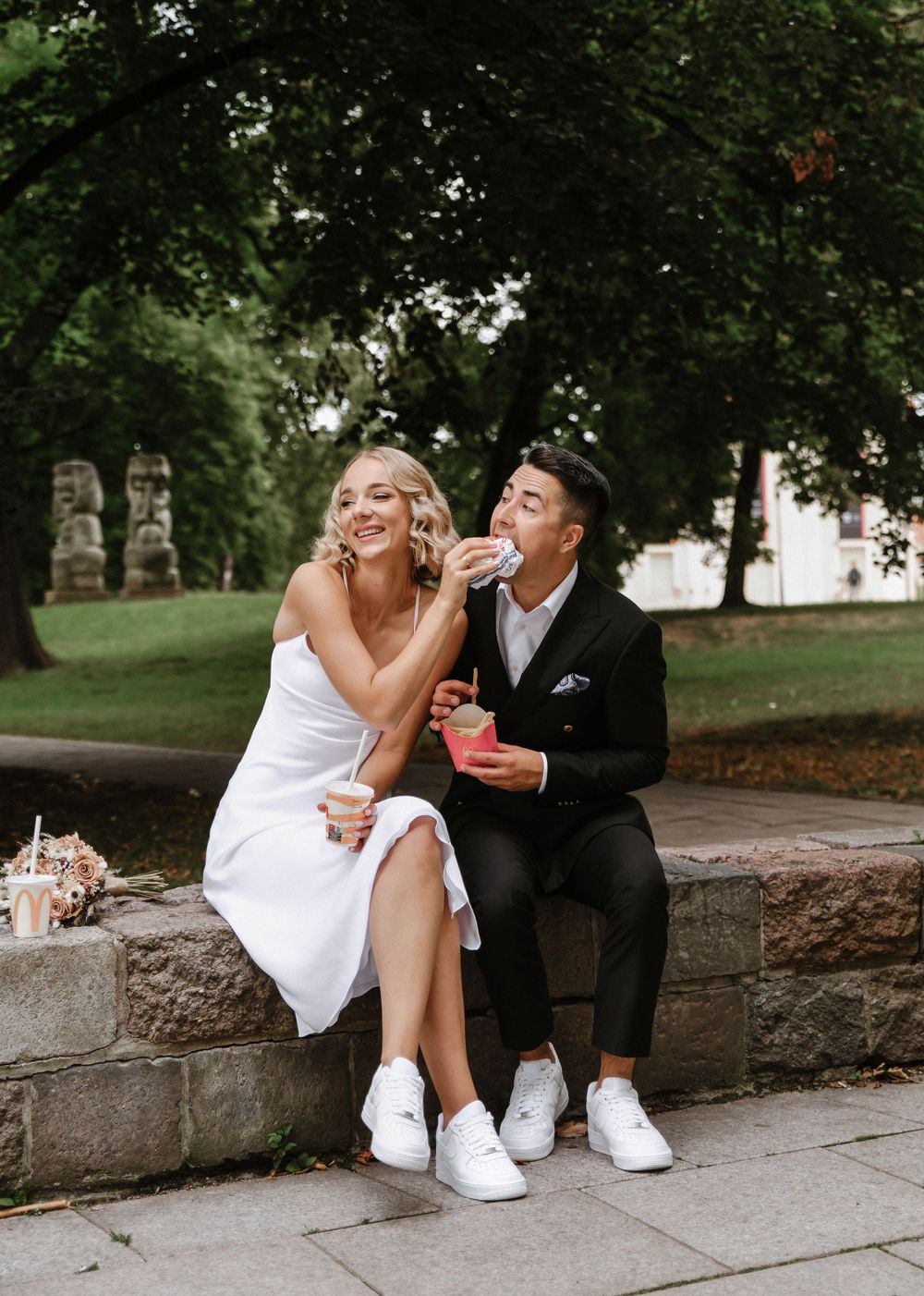 Playful couple in wedding attire sharing snacks while sitting on a stone wall.
