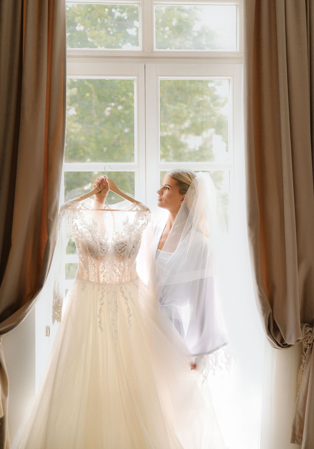 Wedding dresses displayed on hangers by large windows in a bridal suite with soft natural lighting and brown curtains.