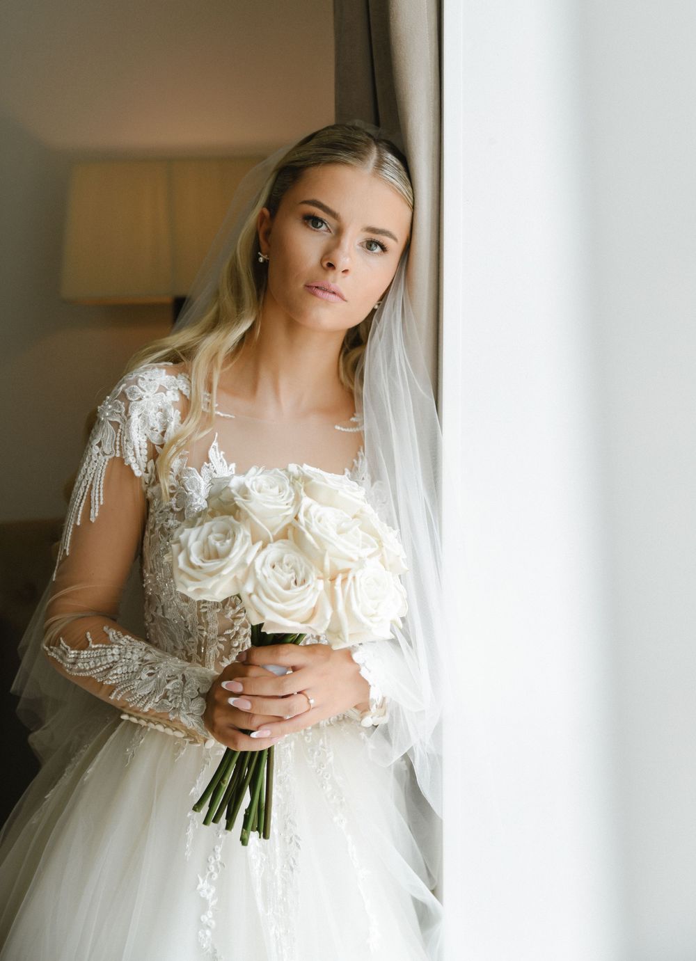 A bride in a delicate lace wedding dress holds a classic white rose bouquet while standing near a window.