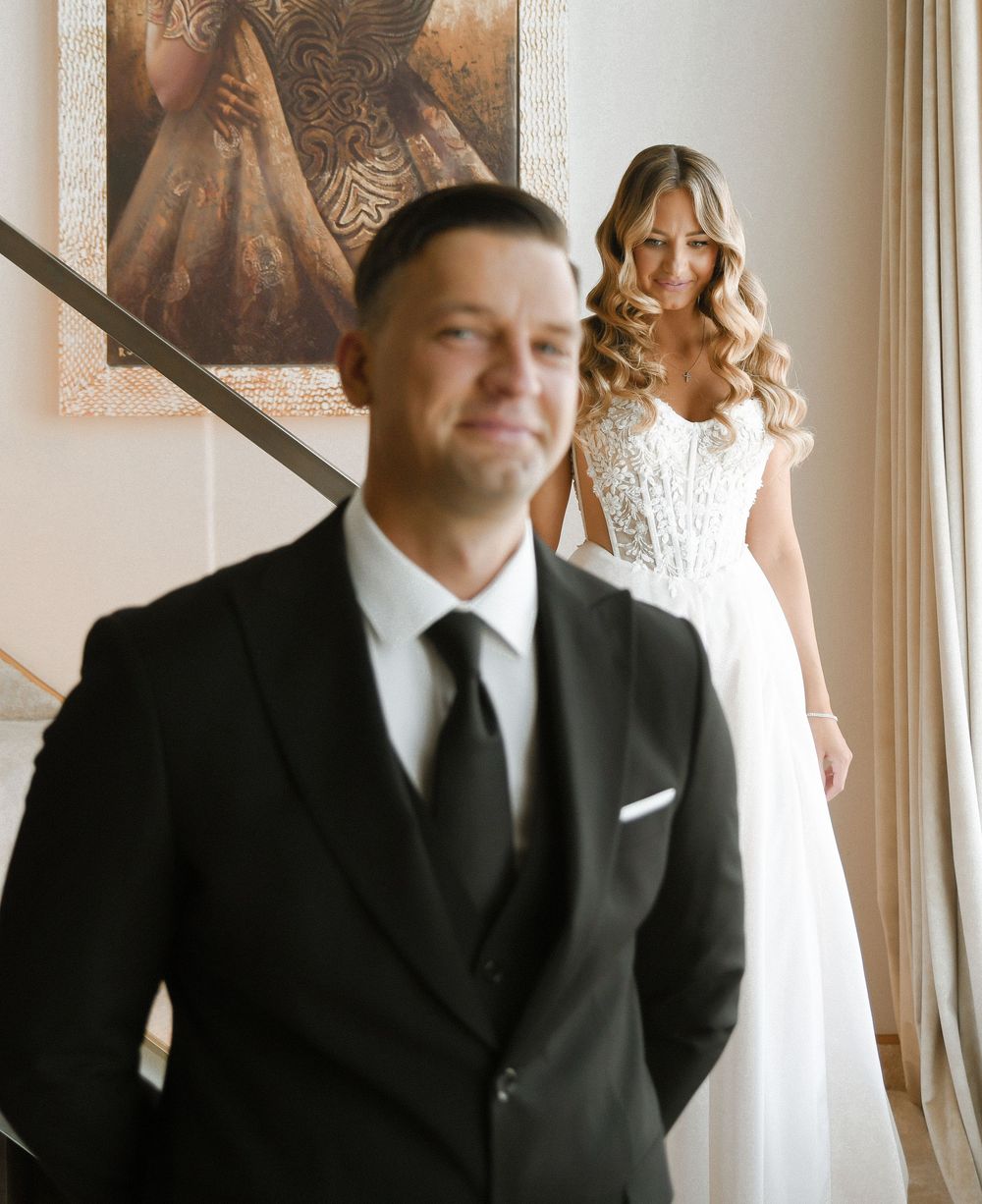Elegant wedding photo showing a groom in classic black suit and bride in white lace dress standing by a window.
